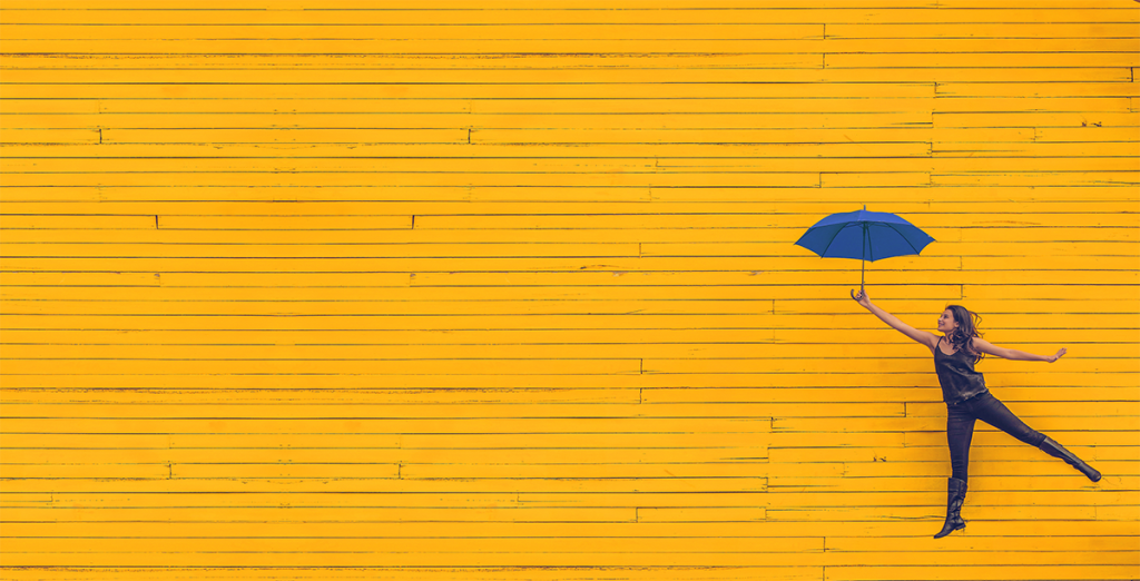 A young women leaping lightly with a blue umbrella in her hand in front of a yellow wall.
