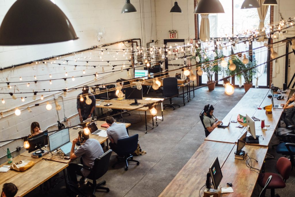 An open office with long wooden tables and office chairs. Above the tables, strings of lights hang from one side of the room to the other. A the front, a set of door length windows with 2 plants. 